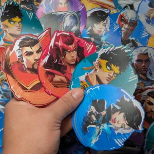 May include: A collection of colourful character stickers featuring various characters with different hairstyles, clothing, and expressions. The stickers are round and have a glossy finish.