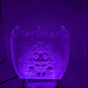 May include: A purple LED night light featuring a cartoon character with a skull-like face, crown, and cape. The design is framed by stylized flames. The night light sits on a black base with a power button.