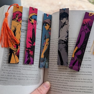 May include: A collection of colorful anime-style bookmarks with tassel accents. Each bookmark features a different character illustration in vibrant hues. The bookmarks are placed on the pages of an open book.