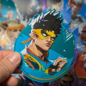 May include: A round sticker featuring a cartoon character with black hair, a yellow headband, and a blue and yellow outfit. The character is wearing a blue mask with a yellow stripe. The sticker has a blue background with a white outline.