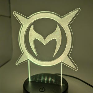 May include: A clear acrylic LED lamp with a stylized emblem. The emblem features a central 'M' shape, encircled by a frame with four pointed extensions. The lamp is on a black base with a power button.