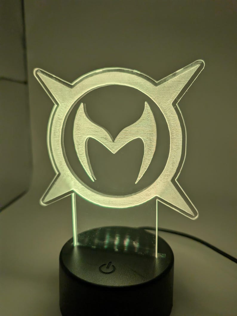 May include: A clear acrylic LED lamp with a stylized emblem. The emblem features a central 'M' shape, encircled by a frame with four pointed extensions. The lamp is on a black base with a power button.