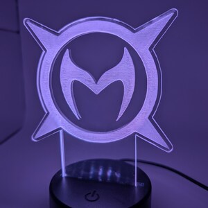 May include: A purple-lit acrylic lamp with a stylized emblem. The emblem is a circular design with a central 'M' shape, surrounded by four pointed shapes. The lamp sits on a black base with a power symbol.