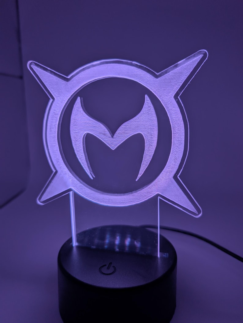 May include: A purple-lit acrylic lamp with a stylized emblem. The emblem is a circular design with a central 'M' shape, surrounded by four pointed shapes. The lamp sits on a black base with a power symbol.