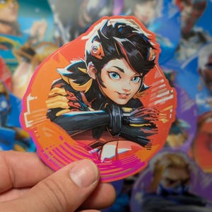 Puede incluir: Una pegatina colorida con una mujer de estilo anime de dibujos animados con cabello negro, ojos azules y un fondo rosa y naranja. La mujer lleva un atuendo amarillo y negro y tiene una expresi&oacute;n decidida en su rostro.