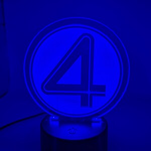 May include: A blue illuminated LED lamp featuring the number 4 inside a circular frame. The lamp is mounted on a black cylindrical base. The design is lit with a bright blue glow.