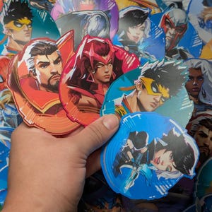 May include: A hand is holding a selection of colourful stickers featuring various characters. The stickers are round and have a glossy finish. The characters are drawn in a cartoon style and include a woman with red hair, a man with a yellow mask, and a woman with blue hair.