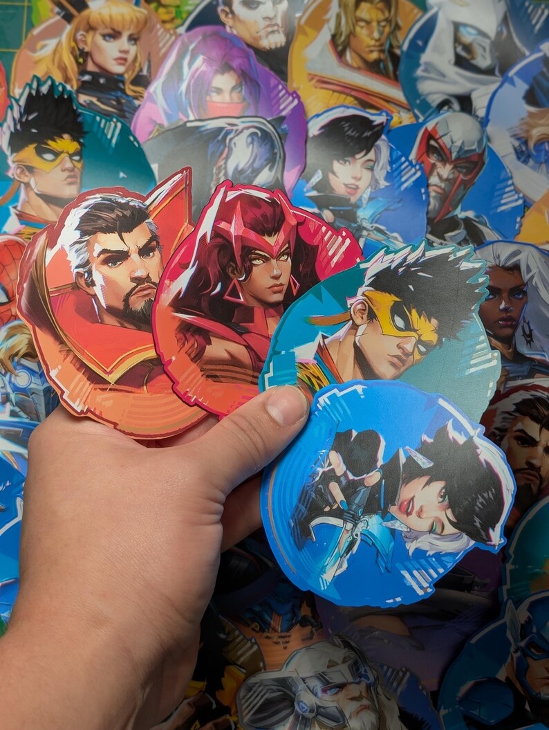 May include: A hand is holding a selection of colourful stickers featuring various characters. The stickers are round and have a glossy finish. The characters are drawn in a cartoon style and include a woman with red hair, a man with a yellow mask, and a woman with blue hair.