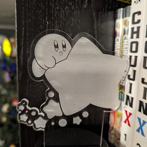 May include: A clear acrylic night light featuring a cartoon character inside a star. The character has large eyes and a small mouth. The light is mounted on a black base with an on/off button. The background includes manga books.