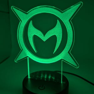May include: A green LED lamp with a distinctive emblem, featuring a central 'M' shape within a circular design, with pointed extensions. The lamp emits a green glow. The black base includes a power button.