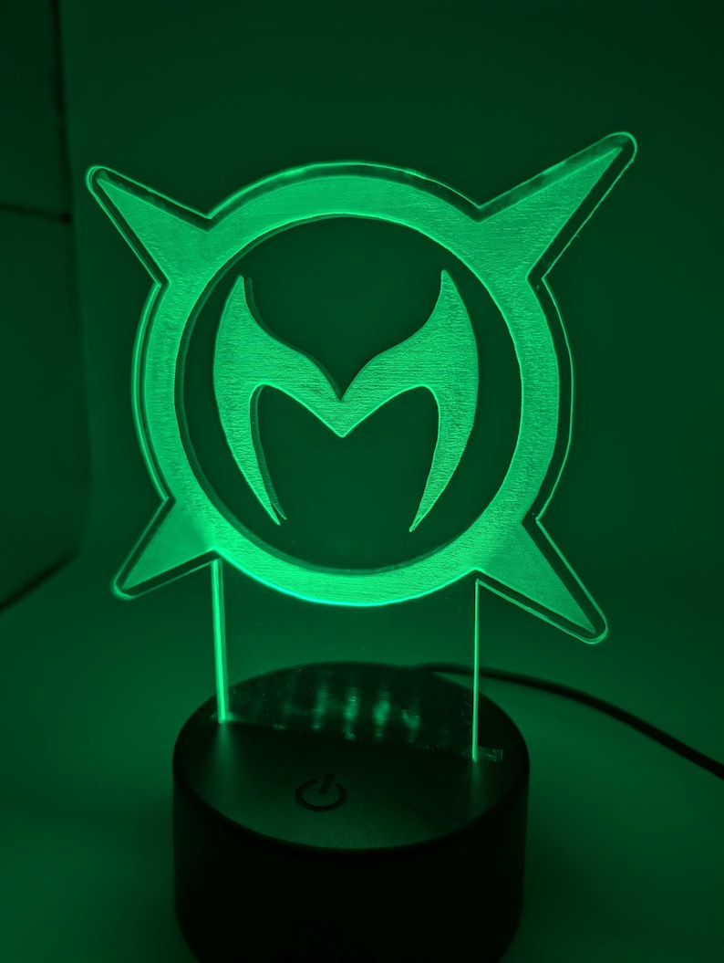 May include: A green LED lamp with a distinctive emblem, featuring a central 'M' shape within a circular design, with pointed extensions. The lamp emits a green glow. The black base includes a power button.