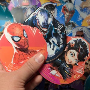 Puede incluir: Tres pegatinas redondas con personajes populares de c&oacute;mics. La primera pegatina presenta a Spider-Man en rojo y azul, la segunda pegatina presenta a Venom en blanco y negro, y la tercera pegatina presenta a un personaje femenino con cabello negro y un atuendo rojo y negro.