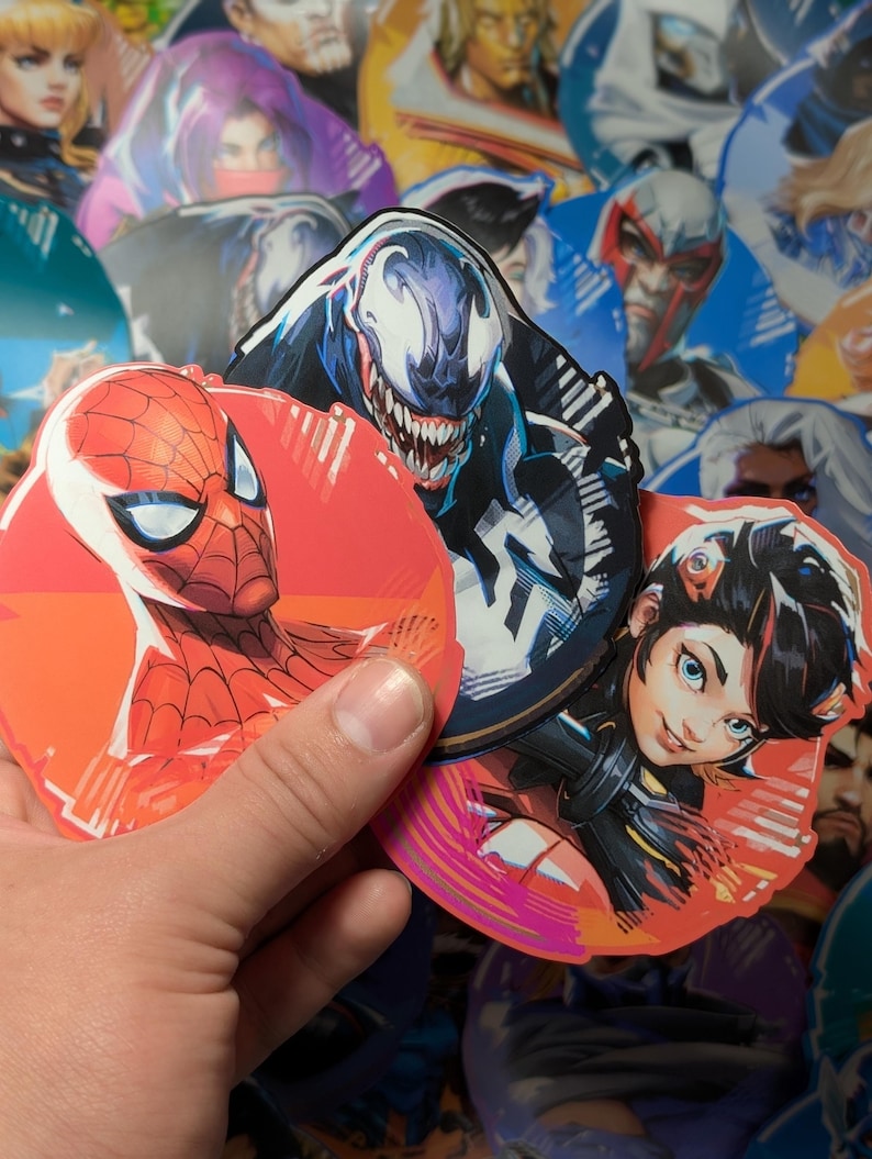 Puede incluir: Tres pegatinas redondas con personajes populares de c&oacute;mics. La primera pegatina presenta a Spider-Man en rojo y azul, la segunda pegatina presenta a Venom en blanco y negro, y la tercera pegatina presenta a un personaje femenino con cabello negro y un atuendo rojo y negro.
