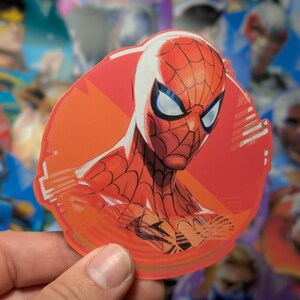 Puede incluir: Una pegatina circular de Spider-Man roja y azul con un contorno blanco. La pegatina tiene un fondo rojo con un dise&ntilde;o blanco y naranja.