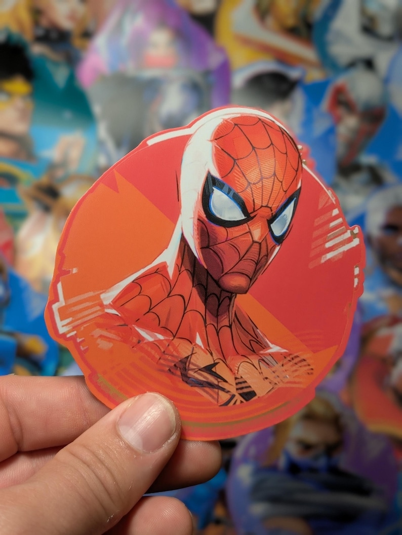 Puede incluir: Una pegatina circular de Spider-Man roja y azul con un contorno blanco. La pegatina tiene un fondo rojo con un dise&ntilde;o blanco y naranja.