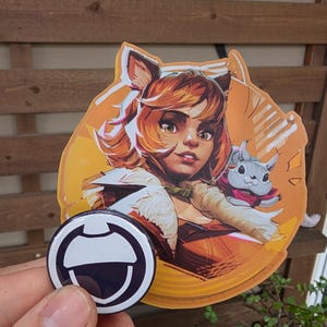 May include: A round, black and white pin with a stylized design is held in front of a large sticker. The sticker features a cartoon illustration of a woman with orange hair and a small gray animal. The background is a wooden fence.