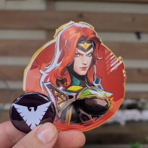May include: A sticker and a button featuring a comic book style illustration of a woman with red hair, green eyes, and a gold headpiece. The button is dark purple with a white bird emblem.