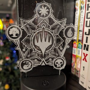 May include: A clear acrylic night light featuring a detailed, geometric design with symbols from Magic: The Gathering. The central emblem is a stylized hand, surrounded by symbols representing different mana colors. The light sits on a black base.
