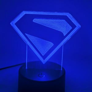 May include: A blue-lit Superman logo night light. The iconic "S" emblem is encased in a diamond shape, illuminated against a dark base. The light has a power button and a cord for plugging into an outlet.