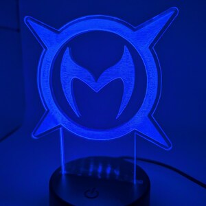 May include: A blue LED lamp displaying a stylized emblem with a central 'M' shape within a circular frame with four pointed extensions. The lamp is on a black base with a power button, and is lit with a bright blue light.