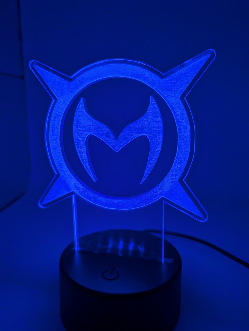 May include: A blue LED lamp displaying a stylized emblem with a central 'M' shape within a circular frame with four pointed extensions. The lamp is on a black base with a power button, and is lit with a bright blue light.