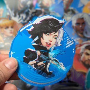 May include: A blue and white circular sticker featuring a cartoon illustration of a woman with black hair and blue eyes. She is wearing a black and white outfit and holding a weapon. The sticker has a white border and a blue background.
