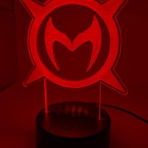 May include: A red LED lamp featuring a stylized emblem with a central 'M' shape within a circular design with four pointed extensions. The lamp sits on a black base with a power button.