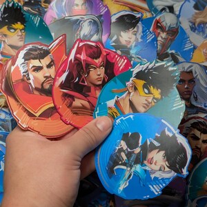 May include: A collection of colourful round stickers featuring various characters from a video game. The stickers are printed on glossy paper and have a holographic finish. The characters are depicted in a variety of poses and expressions, and the stickers are perfect for adding a touch of personality to laptops, water bottles, or other items.