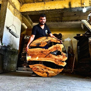 Può includere: Un grande orologio rotondo in legno con numeri romani neri e una lancetta nera. Il quadrante dell'orologio è in legno naturale con resina marrone scuro che riempie gli spazi vuoti. L'orologio è tenuto da una persona in un laboratorio.