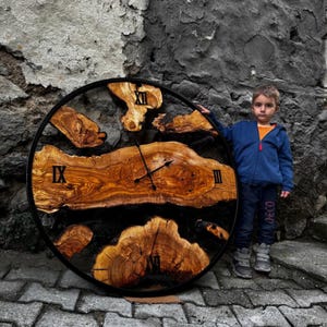 Può includere: Un grande orologio rotondo con numeri romani su un quadrante in legno. L'orologio è in legno con accenti di resina nera e ha una cornice in metallo nero. Un giovane sta in piedi accanto all'orologio.