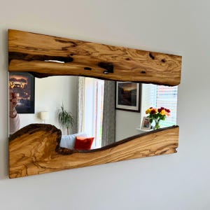 Puede incluir: Un espejo rectangular con marco de madera natural. El marco es de madera marrón claro con nudos marrones oscuros y un diseño de borde vivo. El espejo refleja una habitación con una ventana y obras de arte.