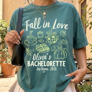 May include: A teal green t-shirt featuring the text Fall in Love, Olivia's BACHELORETTE, and Las Vegas, 2026. The graphic includes line art of a coffee cup, candle, disco ball, plaid blanket, pumpkin, and leaves in a light lime green color.