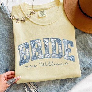 May include: A light yellow t-shirt with the word "BRIDE" in blue floral print lettering. The text "mrs. Williams" is below the word "BRIDE". The shirt is displayed on a pair of blue jeans, with a brown hat and a floral necklace.