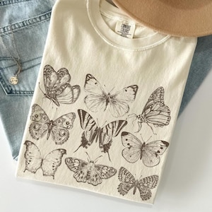 May include: Ivory-colored t-shirt featuring a collection of detailed butterfly illustrations in shades of brown. The shirt is laid on top of light blue denim jeans, with a beige hat and a delicate gold necklace visible.