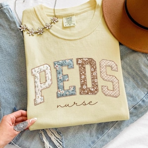 May include: A light yellow t-shirt with the word "PEDS" in patchwork letters, with "nurse" written below. The shirt is laid on top of blue jeans, with a brown hat and a floral necklace. The word "BANANA" is in a circle in the bottom left corner.