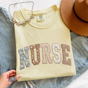 Peut inclure: Un t-shirt jaune clair avec le mot "NURSE" en lettres patchwork. Les lettres sont faites de motifs floraux, à carreaux et unis. Le t-shirt est posé sur un jean bleu, avec un chapeau marron et un collier floral.