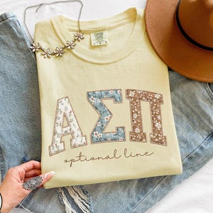 May include: A pale yellow t-shirt featuring the Greek letters ΑΣΠ in floral patchwork. The text "optional line" is below the letters. The shirt is on blue jeans, with a brown hat and a floral necklace.