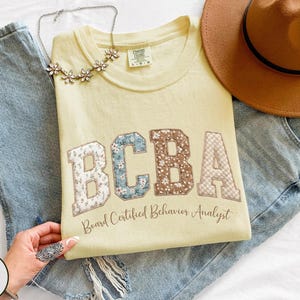 May include: A light yellow sweatshirt with the letters "BCBA" in floral and patterned fabric. The text "Board Certified Behavior Analyst" is below the letters. The sweatshirt is laid on top of blue jeans, with a brown hat and a floral necklace.