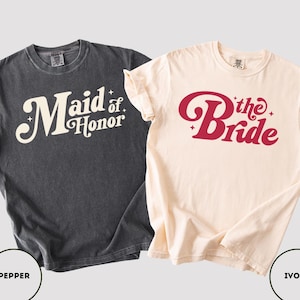 May include: Two t-shirts, one in pepper and one in ivory. The pepper-coloured shirt says "Maid of Honor" in a retro font. The ivory shirt says "The Bride" in a similar font. Both are short-sleeved.