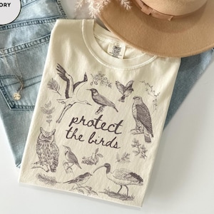 May include: Ivory-colored t-shirt with a graphic of various birds and the text "protect the birds." The shirt is laid on top of light blue denim jeans, with a tan hat and a gold necklace visible in the background.
