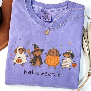 Op de afbeelding: Een violetkleurig T-shirt met korte mouwen en een afbeelding van vier teckels in Halloween-kostuums: een spook, een heks, een pompoen en een mummie. De tekst "halloweenie" staat onder de honden en het shirt heeft een Comfort Colors label.
