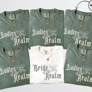 May include: Six t-shirts, five in moss green and one in cream. The green shirts read "Ladies of the Realm" in white script with a dragon graphic. The cream shirt says "Bride of the Realm". Each shirt has a gold necklace.
