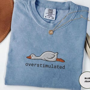 May include: A blue jean-coloured t-shirt with the word "overstimulated" embroidered below a cartoon goose. The shirt has a crew neck and short sleeves. A gold necklace with a pendant is draped across the shirt.