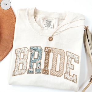 May include: Ivory-colored sweatshirt with the word "BRIDE" in a floral patchwork design. A gold necklace with a pendant rests on the shirt. The shirt is folded and has a tag that says "Comfort Colors".
