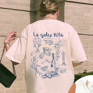 Puede incluir: Una camiseta blanca con un estampado gr&aacute;fico azul de una langosta, uvas, vino y otros alimentos italianos. El texto "La Dolce Vita" est&aacute; impreso en la parte posterior de la camiseta.