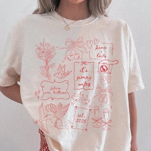 May include: Ivory-colored t-shirt with red graphic illustrations, including flowers, bows, a camera, and text that reads "James + Emily", "It's giving wifey", "future mrs. Williams", and "est. 2025".