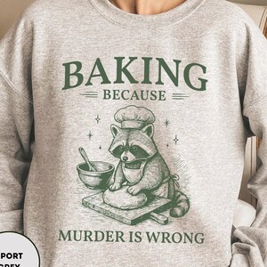 May include: Sport grey sweatshirt with the text "BAKING BECAUSE MURDER IS WRONG". The design features a raccoon chef with a chef's hat and apron, kneading dough on a wooden board. The text and graphic are in green.