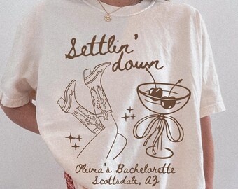May include: Ivory-colored t-shirt with a brown graphic design. The design includes the words "Settlin' down", cowboy boots, a cocktail glass with cherries, and the text "Olivia's Bachelorette Scottsdale, AZ".
