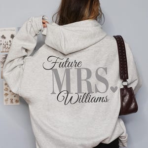May include: Ash gray hoodie with the text "Future MRS Williams" printed on the back. The hoodie has a drawstring hood and a kangaroo pocket. The image also shows a brown purse with a studded strap.