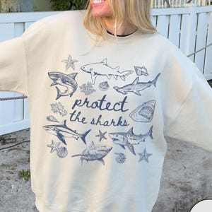 May include: A sand-coloured sweatshirt with a blue graphic of sharks, starfish, and seashells, and the text "protect the sharks." The design features various shark species. A casual, comfortable style.